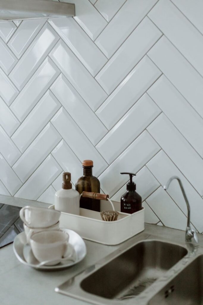 Stylish kitchen sink area with cleaning products and cups on a tiled counter.